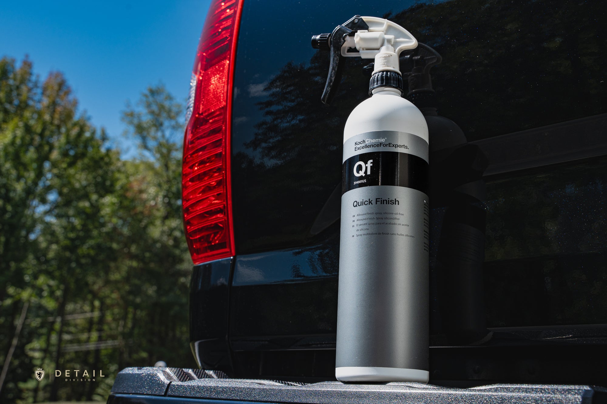 Car detailing spray bottle labeled 'Qf' on a truck tailgate with trees in the background and metal flake popping through black paint